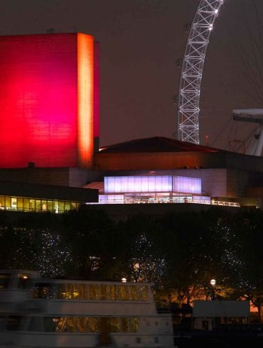 Photo of the National Theatre from the northeast at night-time, with a red-lit flytower and the London Eye