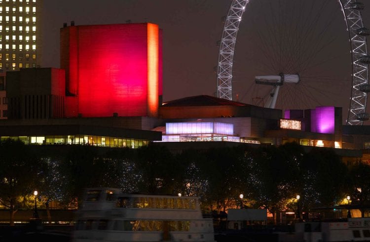 Photo of the National Theatre from the northeast at night-time, with a red-lit flytower and the London Eye