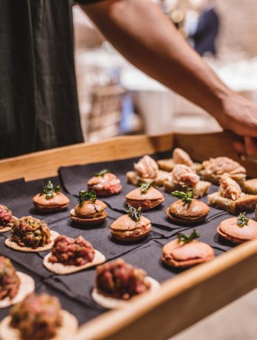 A selection of 3 different types of canapes, 8 of each, on black serviettes in a wooden tray being held by a waiter