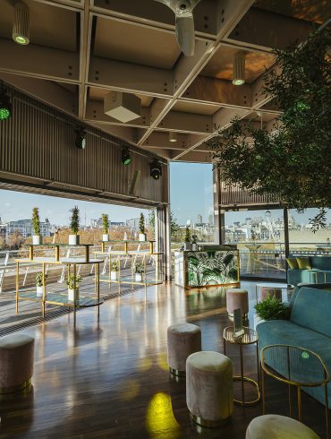 The interior space of the Buffini Chao Deck, with a tree, surrounded by sofas, in the centre of the room., looking out across the London skyl;ine.