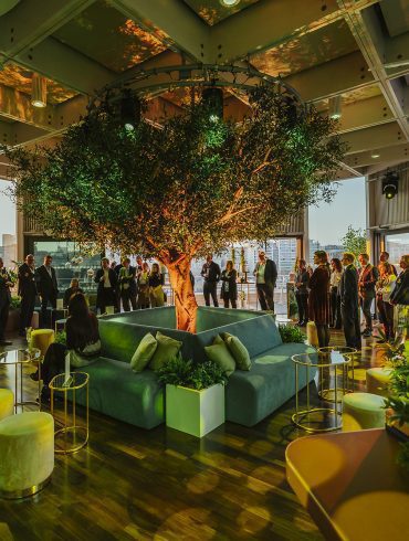 Guests in the interior space of the Buffini Chao Deck, with a tree, surrounded by sofas, in the centre of the room.