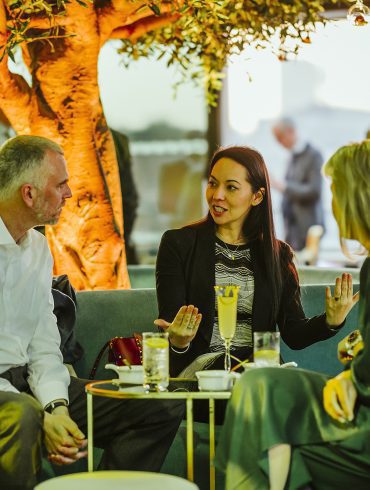 Guests sat on sofas and chairs surrounding a tree, in the interior space of the Buffini Chao Deck.