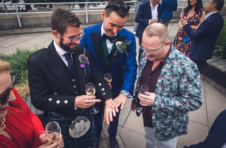 Two grooms with a wedding guest looking at their rings at Buffini Chao Deck